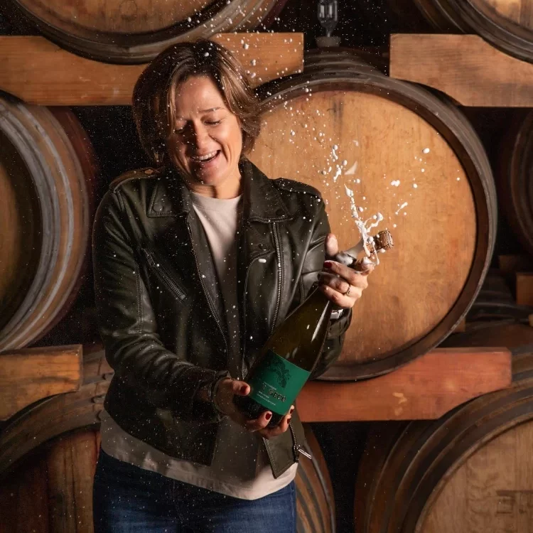 Clarise Sciocatti-Langeveldt, popping a bottle of what looks like sparkling wine standing in front of wine barrels